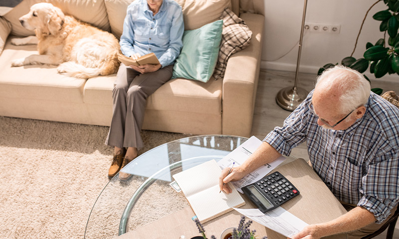 senior man reviewing budget at the kitchen table while his wife reads on the couch with their dog do i have enough to retire